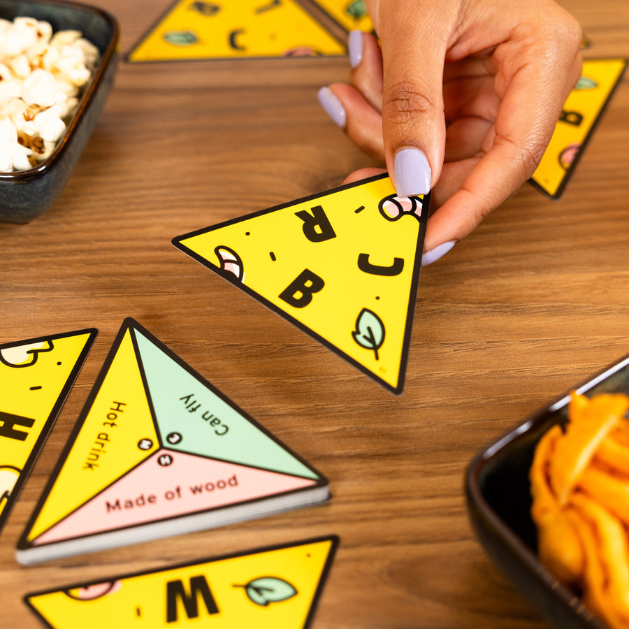 A hand with lavender nail polish holds a yellow triangular card from P for Pizza over a wooden table with other matching cards and snack bowls.