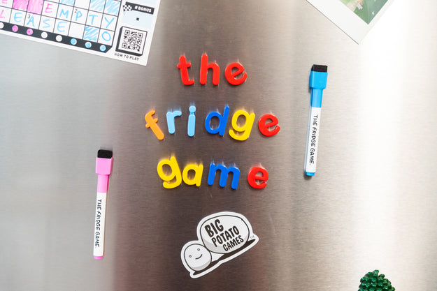 Colorful magnetic letter tiles spelling 'the fridge game' arranged on a stainless steel refrigerator, with two branded magnetic markers and a Big Potato Games sticker visible.